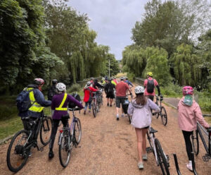A group of cyclists of a variety of ages are seen from behind wheeling their bikes along a tree lined gravel track