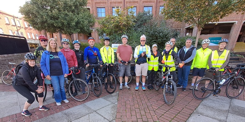 A group of cyclists of various ages and genders assemble outside a red brick building for a ride