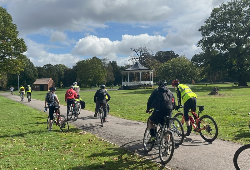 Cyclists ride on a path through a park. A Victorian style covered bandstand is in the background.