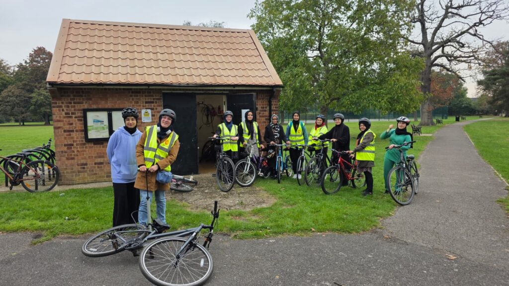 A group of women, some wearing cycle helmets and yellow hi-viz tabards, stand with their bikes in a grassy area in front of a small brick building in a public park. In the foreground are two women standing behind a bicycle which is lying on its side. The woman on the right is operating a track pump.