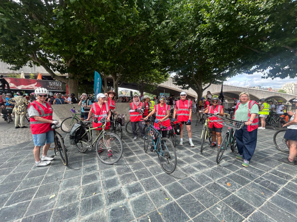 A group of women wearing coral-pink tabards, facing the camera, holding bicycles and smiling. They are in a pedestrianised area and the concrete spans of Waterloo Bridge are in the background.