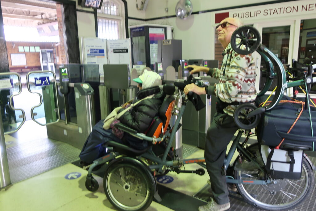 A person sitting on a wheelchair tandem , pedaled by another person, entering Ruislip Metropolitan Line station